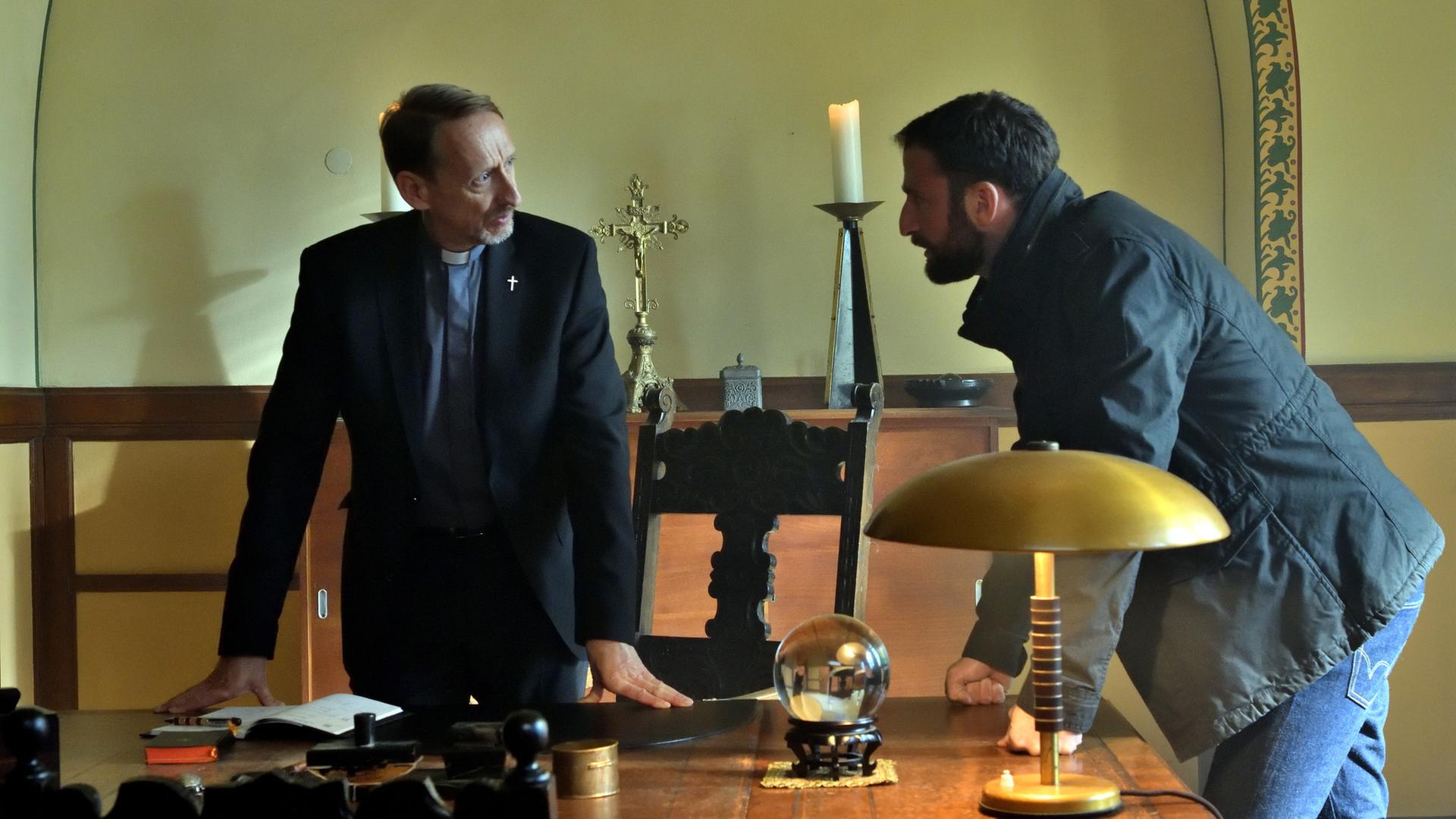 "Die Spezialisten - Die kalte Hand": Im Büro von Pater Thiel (Gilbert von Sohlern) stehen er und Hauptkommissar Henrik Mertens (Matthias Weidenhöfer) nebeneinander. Beide stehen nach vorn übergebeugt und stützen sich mit den Händen auf einem großen Holzschreibtisch ab. Sie sehen sich an.