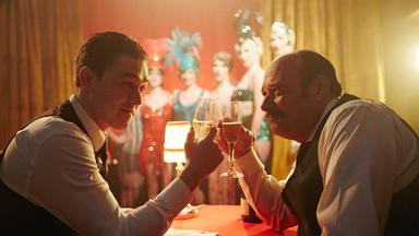 "Die Kanada-Saga - Aufschwung und Krise": Spielszene: Joseph Bisaillon und sein Vorgesetzter, der Chef der kanadischen Zollbehörde, Jacques Bureau, sitzen sich an einem Tisch gegenüber und stoßen mit Sekt an. Im Hintergrund sind unscharf aufreizend gekleidete Damen zu sehen.