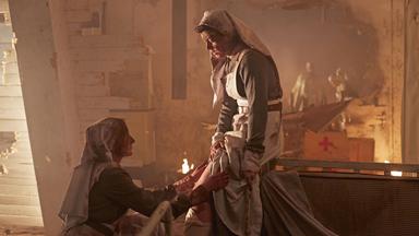 "Die Kanada-Saga - Erster Weltkrieg": Spielszene: Eine Krankenschwester kniet in einem düsteren Raum vor einer Kollegin (beide Darstellerinnnen unbekannt), um deren Bein zu verarzten.