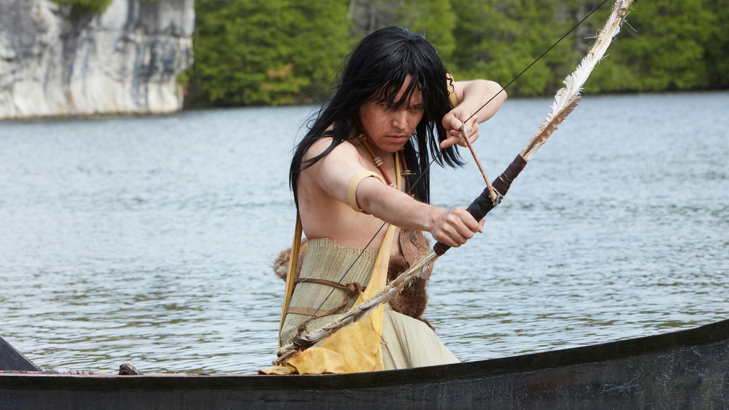 "Die Kanada-Saga - Pioniere und Abenteurer": Spielszene: Häuptling Maquinna (Darsteller unbekannt) vom Volk der Mowachaht sitzt in einem Boot und zielt mit Pfeil und Bogen in Richtung Wasser.