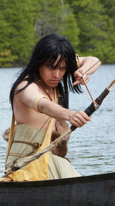 "Die Kanada-Saga - Pioniere und Abenteurer": Spielszene: Häuptling Maquinna (Darsteller unbekannt) vom Volk der Mowachaht sitzt in einem Boot und zielt mit Pfeil und Bogen in Richtung Wasser.