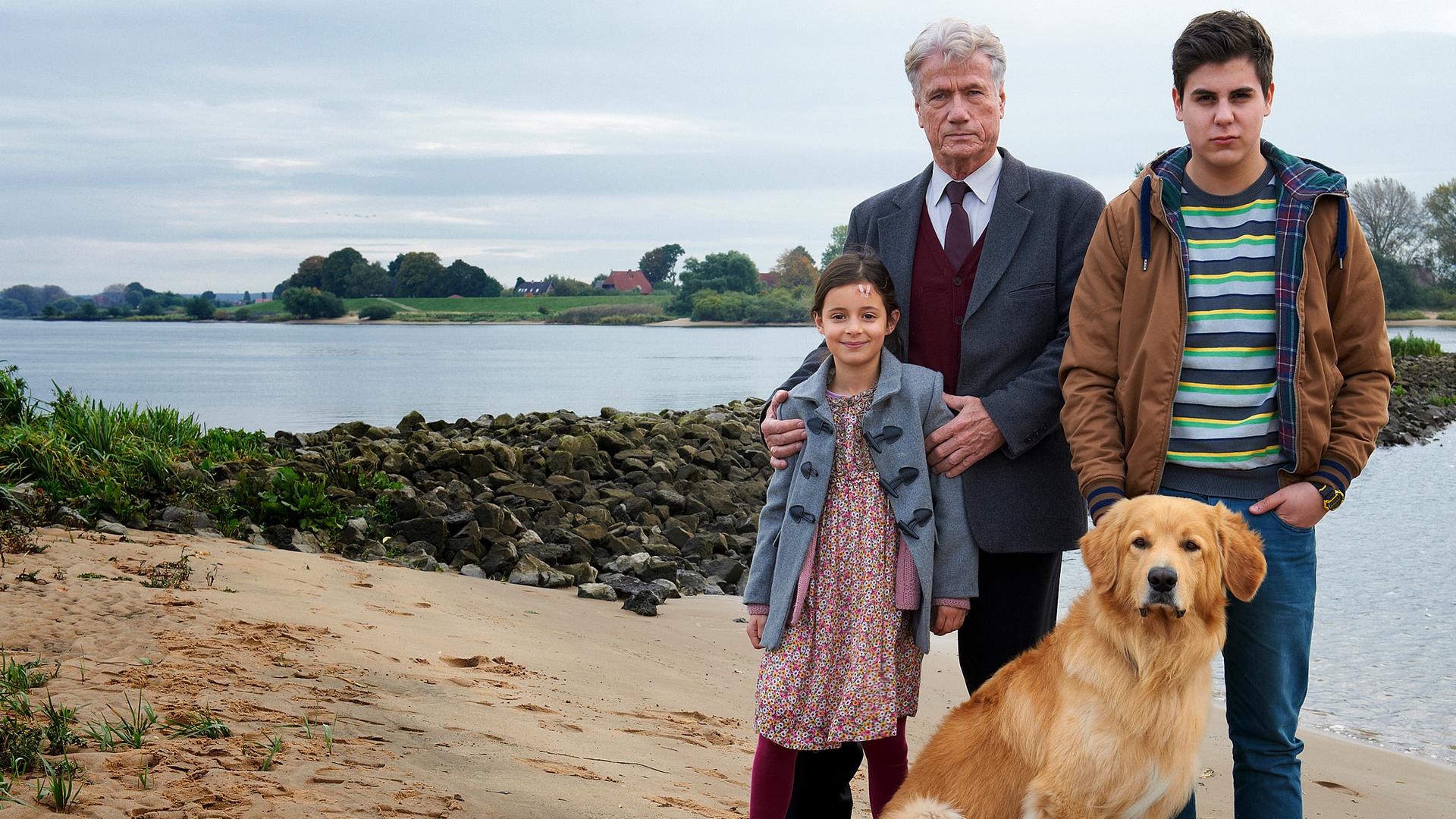 Ernst Blessing (Jürgen Prochnow) steht mit Hewi (Mia Kasalo) und Dilo Amedi (Ugur Ekeroglu) und einem Hund am Strand. Ernst hat seine Hände auf Hewis Schultern gelegt.
