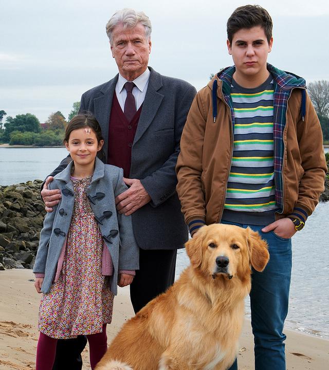 Ernst Blessing (Jürgen Prochnow) steht mit Hewi (Mia Kasalo) und Dilo Amedi (Ugur Ekeroglu) und einem Hund am Strand. Ernst hat seine Hände auf Hewis Schultern gelegt.