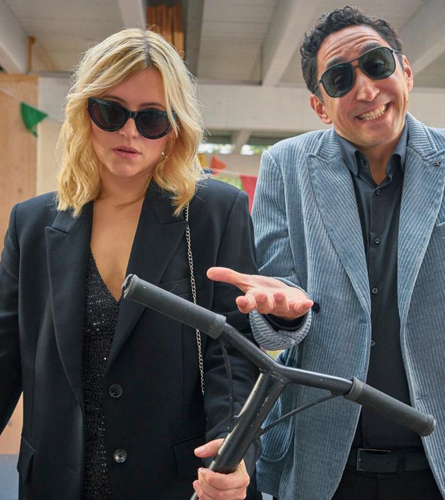 "Die Kinderschwindlerin": Nina (Laura Storz) und ihr Freund Sammy (Ekrem "Eko Fresh" Bora) stehen auf einer bunt geschmückten Veranda. Nina schaut genervt, während Sammy lacht.