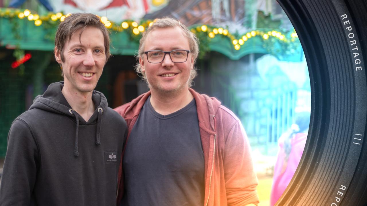 "ZDF.reportage: Die Kirmes Anfänger - Geisterbahn mit Startproblemen": Aiko Bartsch und Stefan Bartsch stehen nebeneinander und halten sich im Arm. Beide lächeln in die Kamera. Im Hintergrund steht ihre Geisterbahn.