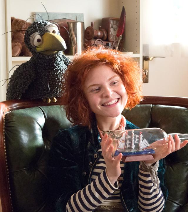 Rudi hockt auf der Lehne eines Sessels, auf dem Klabauterfrau Karli sitzt und ein Buddelschiff in den Händen hält. Beide schauen frech in den Raum.