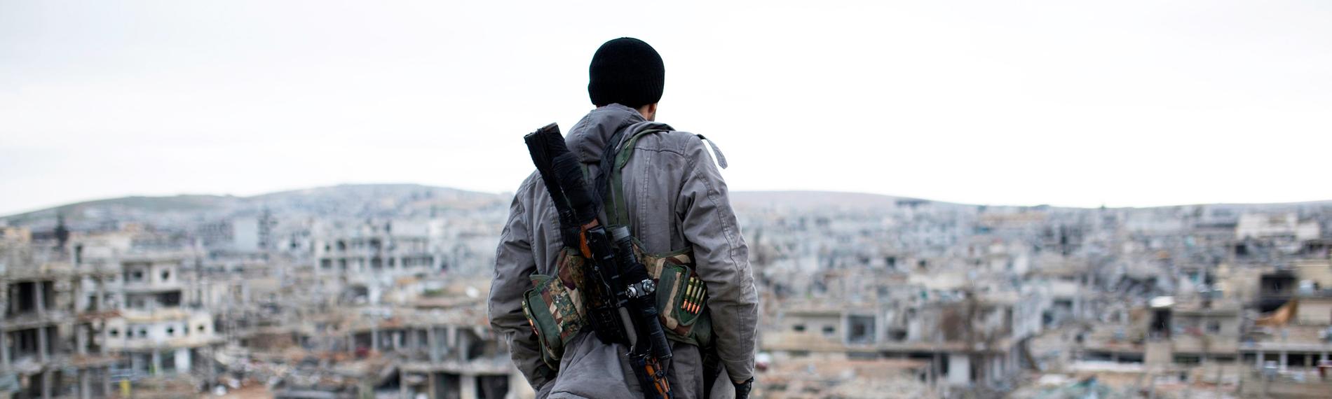 "Die Kurden – Unterdrückung, Terrorismus und Verrat": Ein bewaffneter kurdischer Kämpfer steht mit dem Rücken zur Kamera am Rand eines zerstörten Hauses und schaut auf die ebenfalls zerstörte Stadt vor ihm.