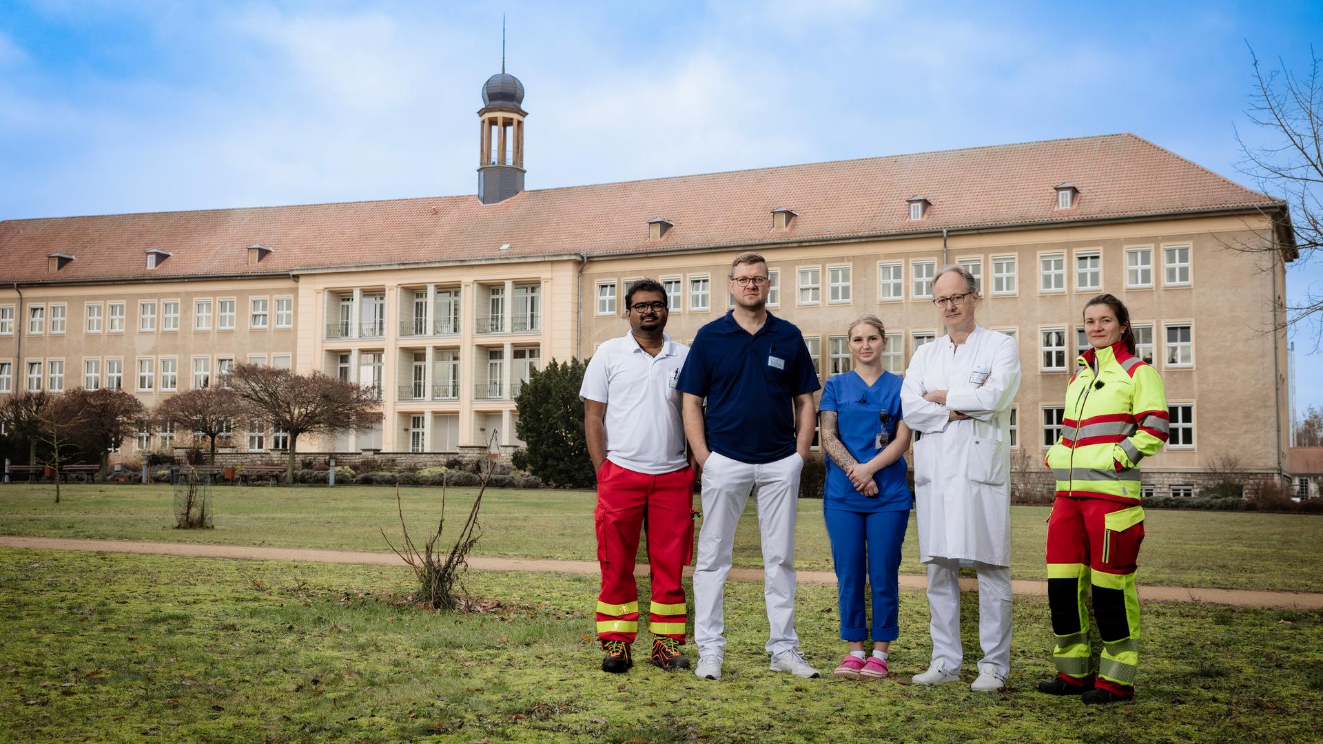Fünf Menschen in medizinischer Kleidung stehen auf einer Wiese vor einem beigefarbenen Krankenhaus-Gebäude: Anoop Puritipati (35, Assistenzarzt), Kai Wilke (42, Orthopäde und Unfallchirurg), Janina Buhe (24, Krankenpflegerin), Georg Bauer (58, Chefarzt) und Julia Kraushaar (35, Assistenzärztin).