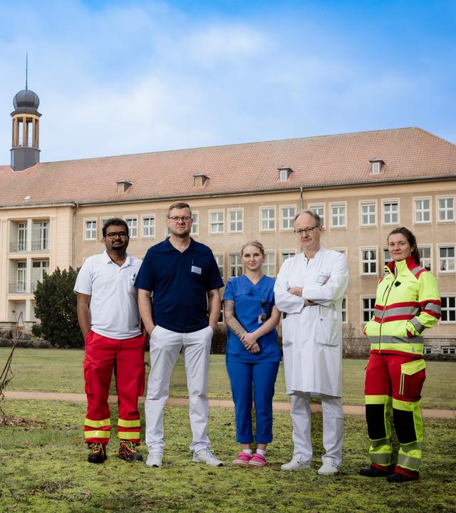 Fünf Menschen in medizinischer Kleidung stehen auf einer Wiese vor einem beigefarbenen Krankenhaus-Gebäude: Anoop Puritipati (35, Assistenzarzt), Kai Wilke (42, Orthopäde und Unfallchirurg), Janina Buhe (24, Krankenpflegerin), Georg Bauer (58, Chefarzt) und Julia Kraushaar (35, Assistenzärztin).