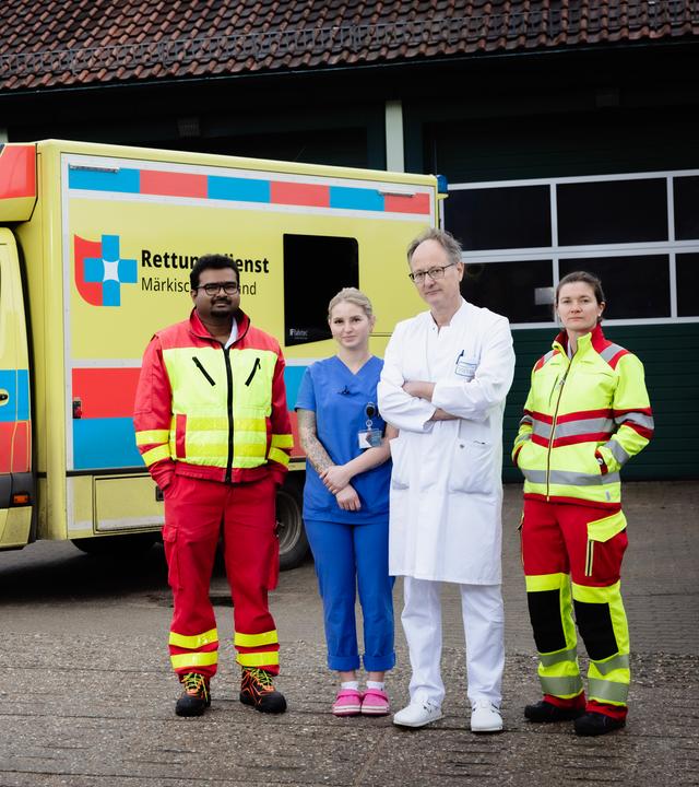 Vier Personen stehen vor einem gelben Rettungswagen mit rot-blauen Quadraten und der Aufschrift "Rettungsdienst Märkisch-Oderland": Anoop Puritipati (35, Assistenzarzt), Janina Buhe (24, Krankenpflegerin), Georg Bauer (58, Chefarzt) und Julia Kraushaar (35, Assistenzärztin).