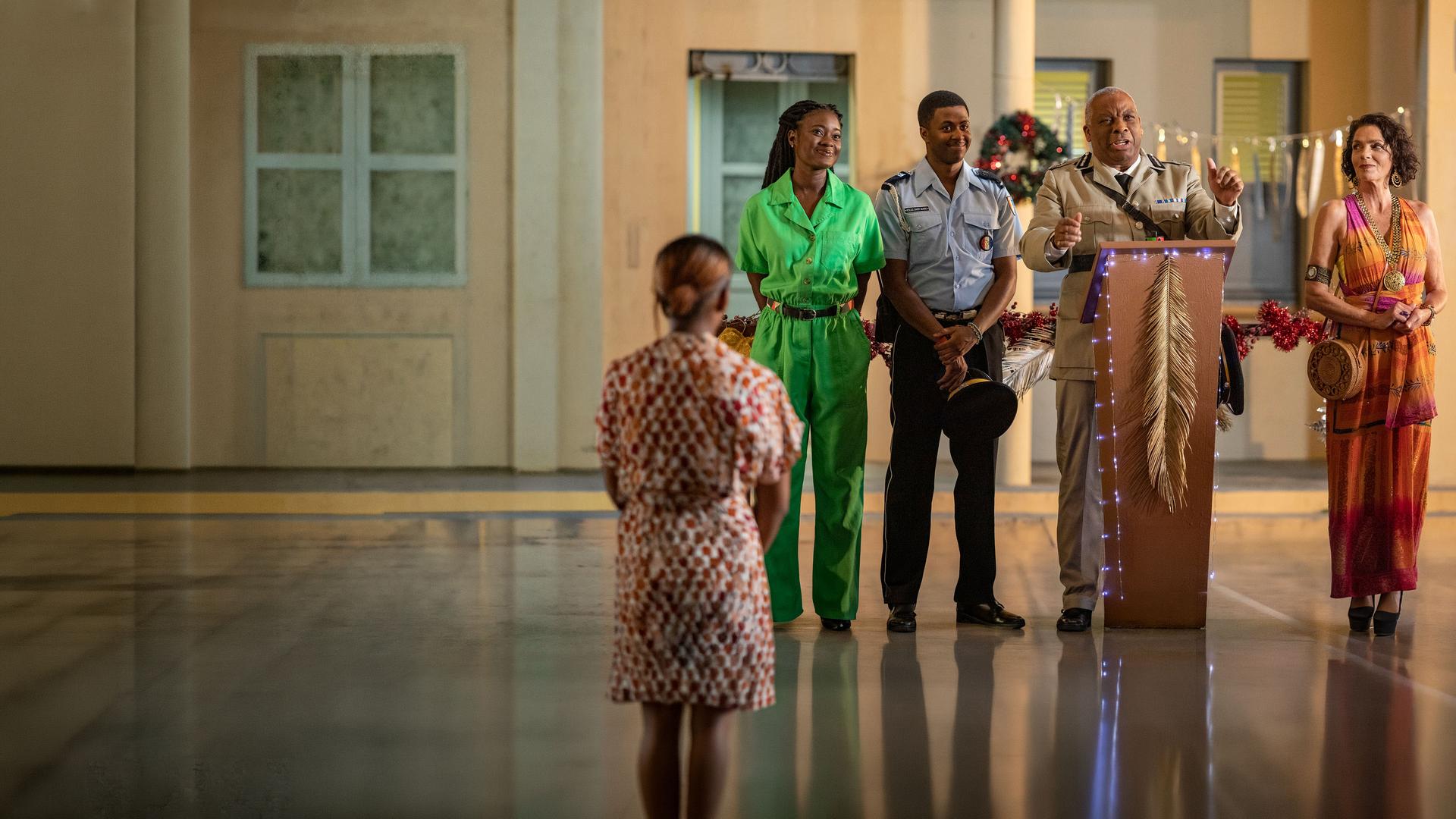"Death in Paradise - Die letzte Botschaft (1)": Commissioner Selwyn Patterson (Don Warrington) steht an einem festlich geschmückten Rednerpult vor einem Gebäude. An seiner Seite stehen Marlon Pryce (Tahj Miles) und Naomi Thomas (Shantol Jackson), die lächelnd geradeaus blicken, sowie Catherine Bordey (Elizabeth Bourgine), die erwartungsvoll zum Commissioner schaut.