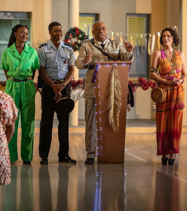 "Death in Paradise - Die letzte Botschaft (1)": Commissioner Selwyn Patterson (Don Warrington) steht an einem festlich geschmückten Rednerpult vor einem Gebäude. An seiner Seite stehen Marlon Pryce (Tahj Miles) und Naomi Thomas (Shantol Jackson), die lächelnd geradeaus blicken, sowie Catherine Bordey (Elizabeth Bourgine), die erwartungsvoll zum Commissioner schaut.