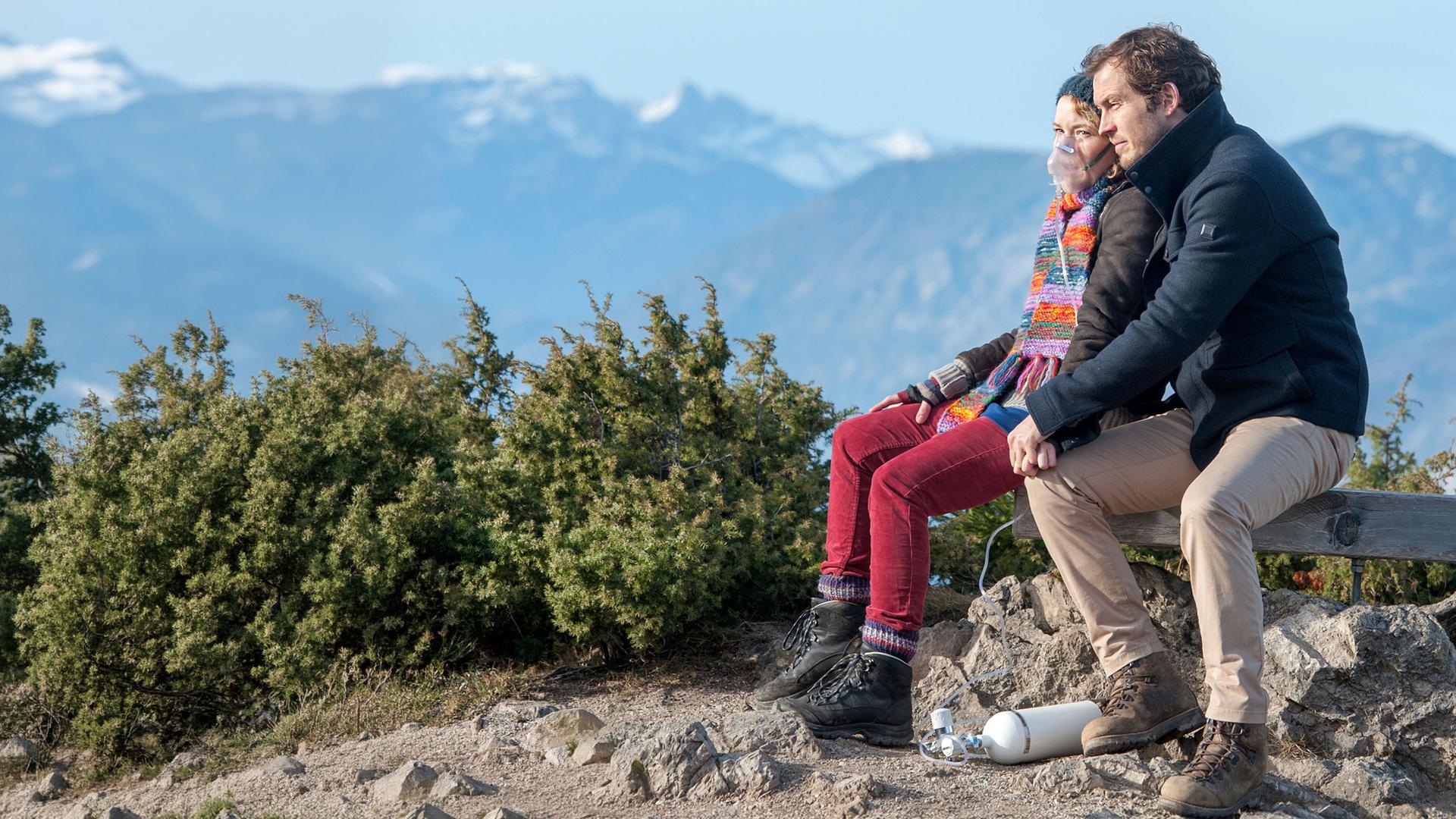 "Der Bergdoktor - Die letzte Reise (2)": Pia (Angelina Häntsch) und Jan (Stefan Murr) sitzen Hand in Hand auf einer Holzbank auf einer Anhöhe, hinter ihnen das Bergpanorama. Pia trägt eine Atemmaske, an die eine Sauerstoffflasche angeschlossen ist, die auf dem Boden zwischen den beiden liegt.