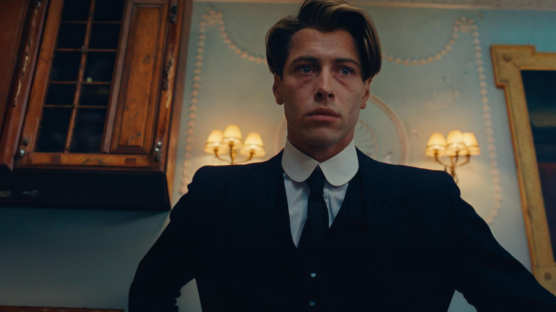 "This Is Not a Murder Mystery - Die Liebenden": Leichte Untersicht, Porträtfoto: René Magritte (Pierre Gervais) befindet sich in einem Raum. Hinter ihm an der Wand hängen große gerahmte Gemälde und Schirmlampen.