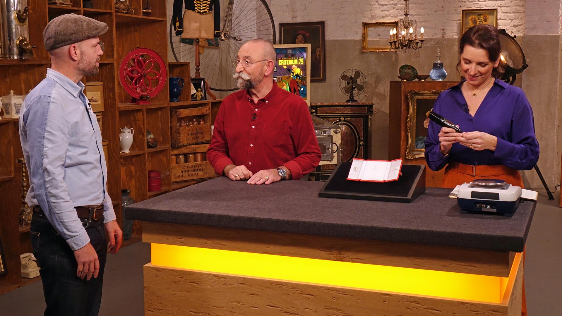 Im Expertisenraum steht der Verkäufer eines von Salvador Dalí entworfenen Kreuzanhängers und spricht mit Horst Lichter. Wendela Horz begutachtet derweil den Anhänger.   