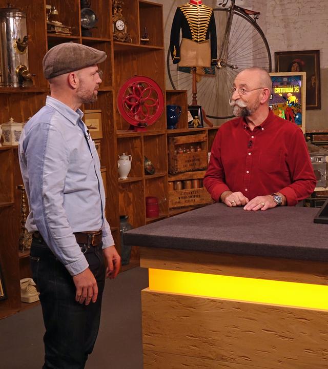 Im Expertisenraum steht der Verkäufer eines von Salvador Dalí entworfenen Kreuzanhängers und spricht mit Horst Lichter. Wendela Horz begutachtet derweil den Anhänger.   