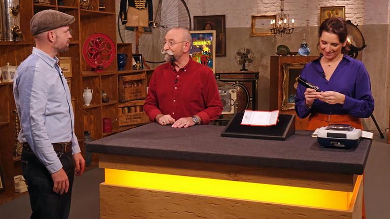 Im Expertisenraum steht der Verkäufer eines von Salvador Dalí entworfenen Kreuzanhängers und spricht mit Horst Lichter. Wendela Horz begutachtet derweil den Anhänger.   