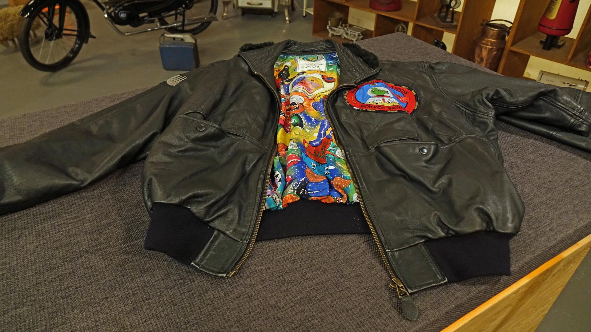 Schwarze Herren-Lederjacke mit buntem Innenfutter in Pop Art-Design. Auf der linken Brust ist ein runder Aufnäher mit einem Hundemotiv angebracht.