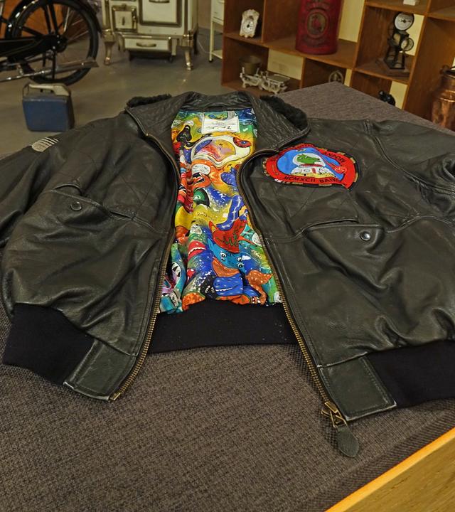 Schwarze Herren-Lederjacke mit buntem Innenfutter in Pop Art-Design. Auf der linken Brust ist ein runder Aufnäher mit einem Hundemotiv angebracht.