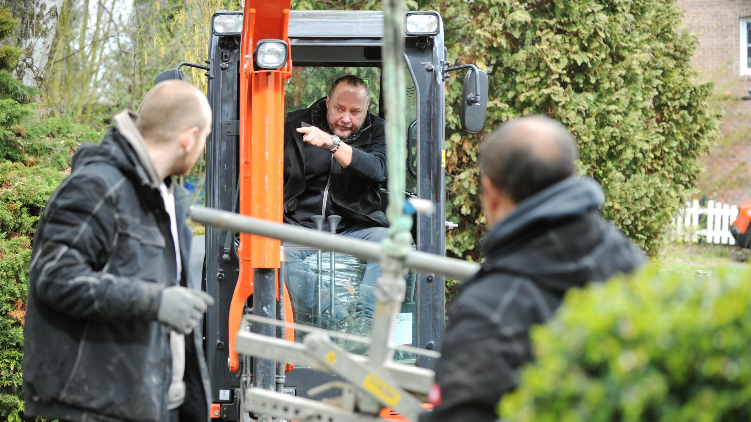Ein Mann sitzt in einem Baufahrzeug. Links und rechts davon stehen zwei Männer, die ihn anschauen.