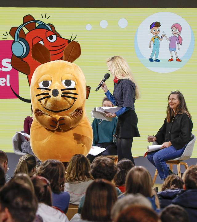 Auf einer Bühne sitzen und stehen mehrere Personen und eine große Maus-Plüschfigur.