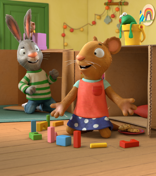 Pip und Posy sind im Kinderzimmer und knien jeweils vor einem Pappkarton. Vor Posy liegen bunte Bauklötze auf dem Boden. Beide unterhalten sich. Im Hintergrund sieht man eine kleine Kommode mit einem CD-Spieler.