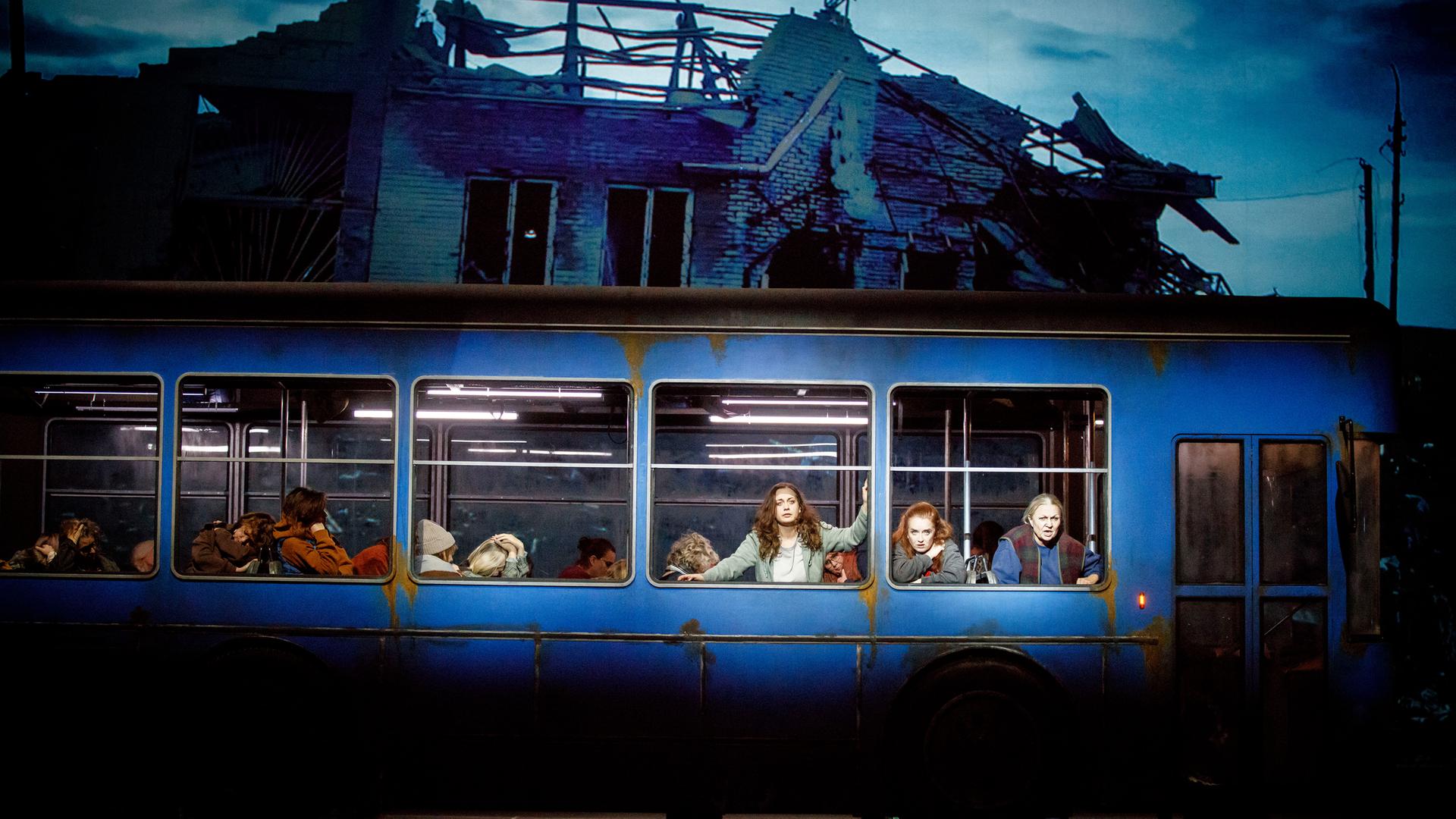 Ein Bus steht auf einer Bühne. Mehrere Darstellerinnen schauen aus dem Bus heraus.Im Hintergrund ist die Ruine eines Hauses zu sehen.