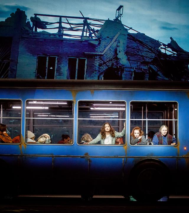 Ein Bus steht auf einer Bühne. Mehrere Darstellerinnen schauen aus dem Bus heraus.Im Hintergrund ist die Ruine eines Hauses zu sehen.