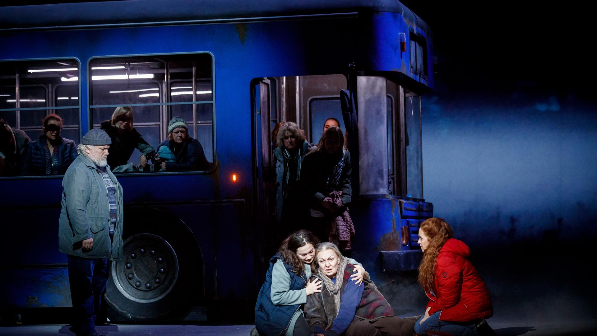 Ein Bus steht auf einer Bühne. Mehrere Darstellerinnen sitzen davor auf dem Boden, während andere Personen noch im Bus sind, oder aussteigen.