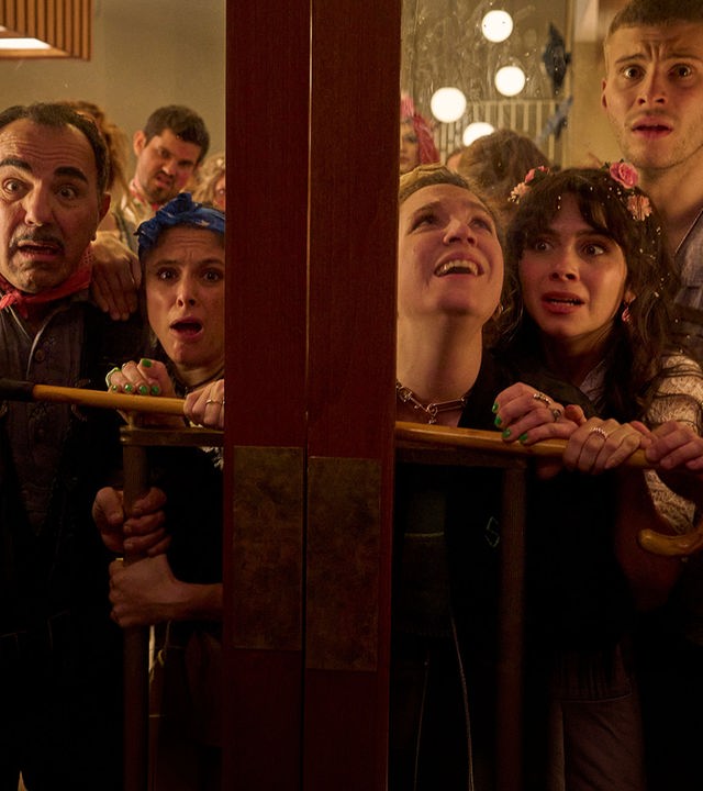 "Nighties - Die Nacht der trinkenden Toten": Die Hotelcrew, Concierge Charles (Adnan Maral), Küchenchefin Franca (Mariananda Schempp), Securitychefin Adriana (Stella Goritzki), Hotelchefin Millie (Tamara Romera Ginés) und Barkeeper Rafael (Ben Felipe), stehen dicht gedrängt hinter einer Glas-Doppeltür. Sie schauen durch die Schreibe und wirken erschrocken. Security-Chefin Adriana hält einen Spazierstock quer gegen die Tür gedrückt.