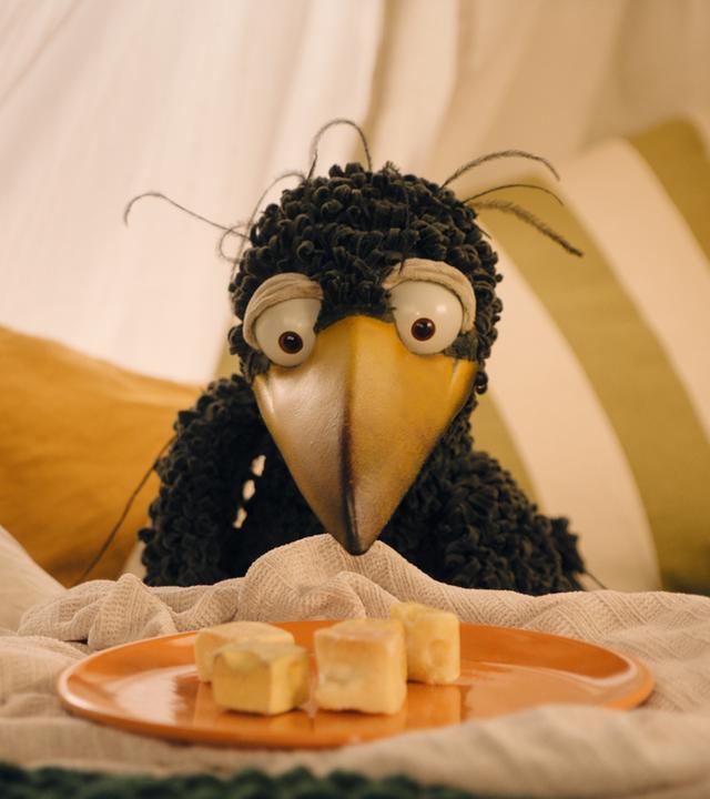 Rabe Rudi sitzt in der gemütlichen Kuschelecke. Vor ihm steht ein orangefarbener Teller, auf dem vier Käsewürfel liegen.