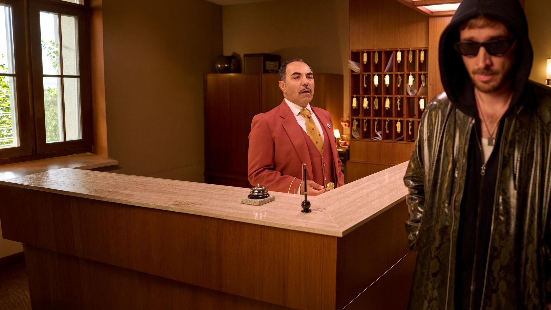 "Nighties - Die Nacht mit dem VIP": Ein Ryan Gosling Double (Tim Vanwersch) läuft in Lederjacke, Kapuzenpullover und Sonnenbrille an einer Hotelrezeption vorbei, hinter der Hotelrezeption steht Charles (Adnan Maral). Hinter ihm steht ein Schlüsselregal, auf dem Tresen liegt eine Klingel und ein Halter mit Stift.