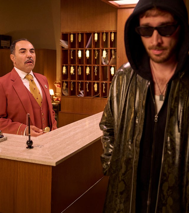 "Nighties - Die Nacht mit dem VIP": Ein Ryan Gosling Double (Tim Vanwersch) läuft in Lederjacke, Kapuzenpullover und Sonnenbrille an einer Hotelrezeption vorbei, hinter der Hotelrezeption steht Charles (Adnan Maral). Hinter ihm steht ein Schlüsselregal, auf dem Tresen liegt eine Klingel und ein Halter mit Stift.