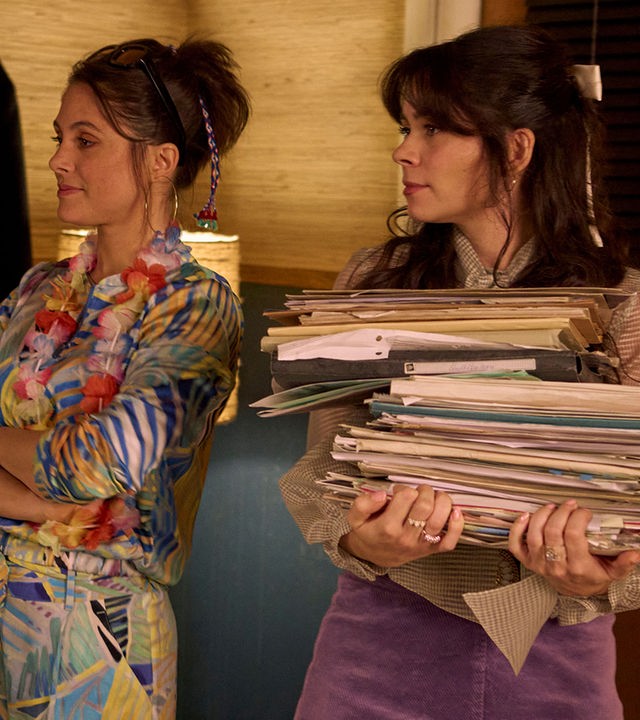 "Nighties - Die Nacht vor der letzten Nacht des Jahres": In einem Innenraum: Franca (Mariananda Schempp) steht mit verschränkten Armen neben Millie (Tamara Romera Ginés), die einen Stapel alter Zeitschriften trägt. Beide schauen lächelnd zur Seite.
