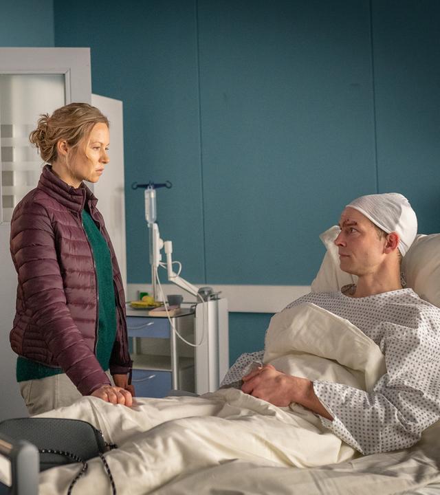 "Notruf Hafenkante - Die Nixe": Jakob Manthey (Christian Clauß) liegt in einem Krankenbett in einem Krankenzimmer im EKH. Sein Kopf ist verbunden. Er blickt zu seiner Frau Nikola (Christin Heim). Sie steht auf der Seite seines Bettes. Der Polizist Mattes Seeler (Matthias Schloo) und seine Kollegin Melanie Hansen (Sanna Englund) stehen nebeneinander hinter Nikola. Beide sehen Jakob Manthey an.