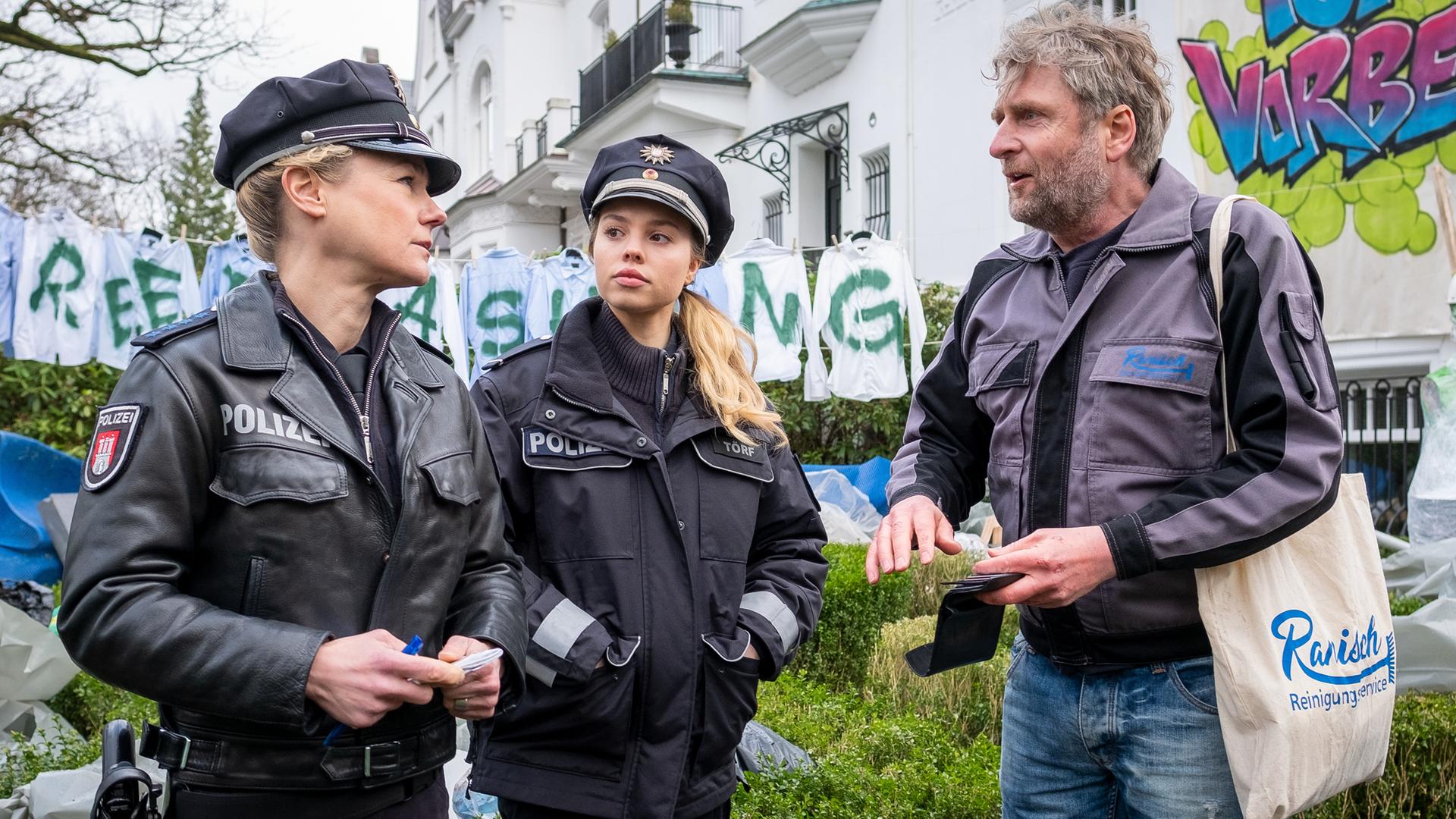 Die Polizistinnen Franzi Jung (Rhea Harder-Vennewald) und Maje Törf (Lea Zoë Voss) stehen nebeneinander auf einem Bürgersteig. Ihnen gegenüber steht Jochen Döring (Jörg Witte). Er ist dabei, etwas aus seinem Geldbeutel zu nehmen. Sie stehen vor dem Vorgarten eines Hauses, in dem verschieden große Gegenstände verpackt rumliegen. 