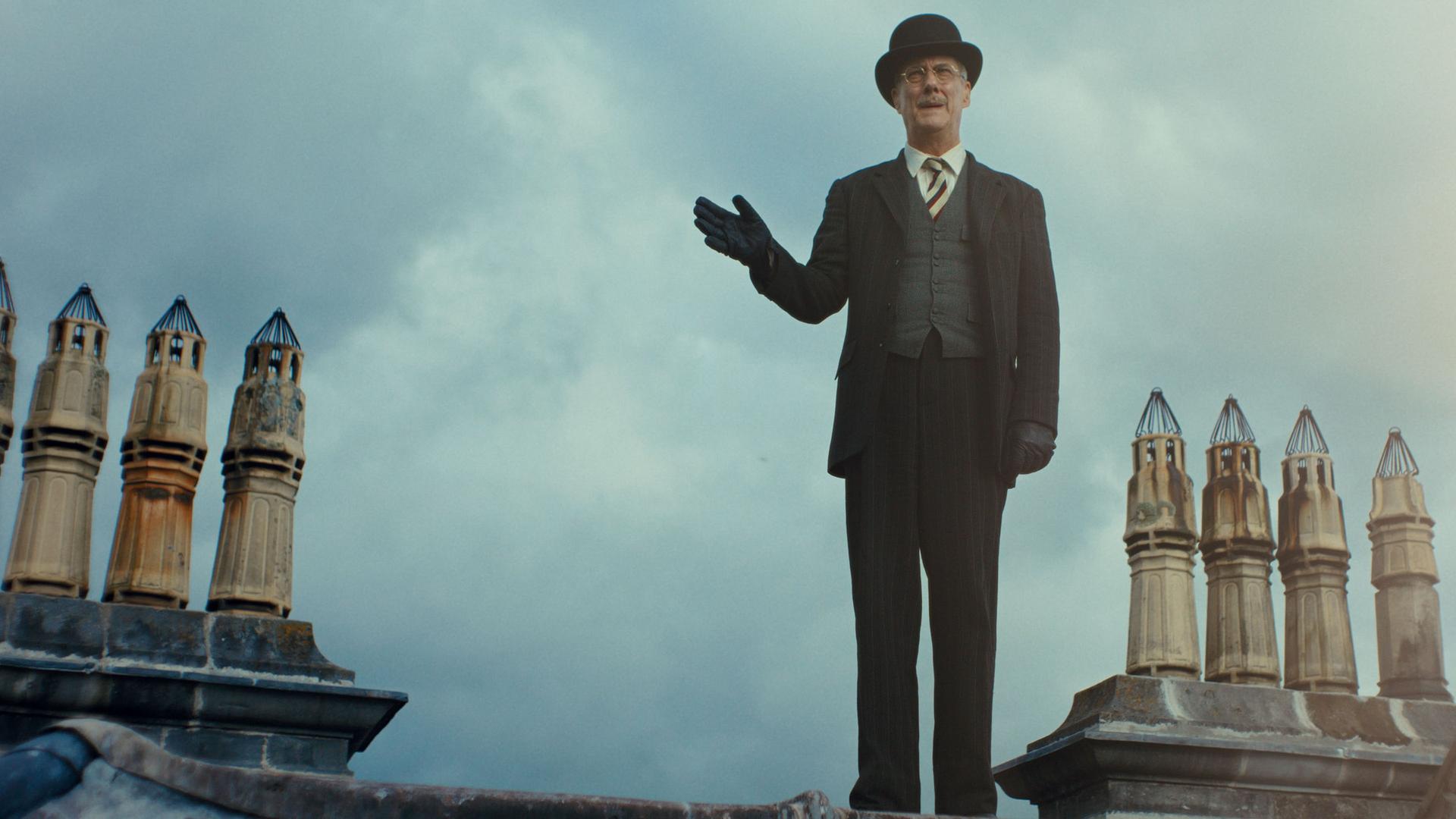 "This Is Not a Murder Mystery - Die Phantomlady": DCI John Thistlethwaite (Stephen Tompkinson) steht auf einem Dachfirst. Neben von ihm ragen mehrere kleine Schornsteine in den Himmel. Er trägt einen Anzug mit Krawatte, Handschuhe und eine Melone. Mit der rechten Hand deutet er zur Seite.