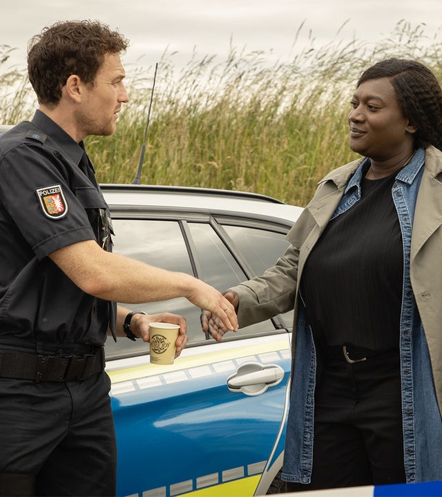 "Die Polizistin und die Sprache des Todes": Pieper Olsen (Artjom Gilz) und Gloria Acheampong (Thelma Buabeng) stehen neben einem Polizeiwagen und geben sich die Hand.