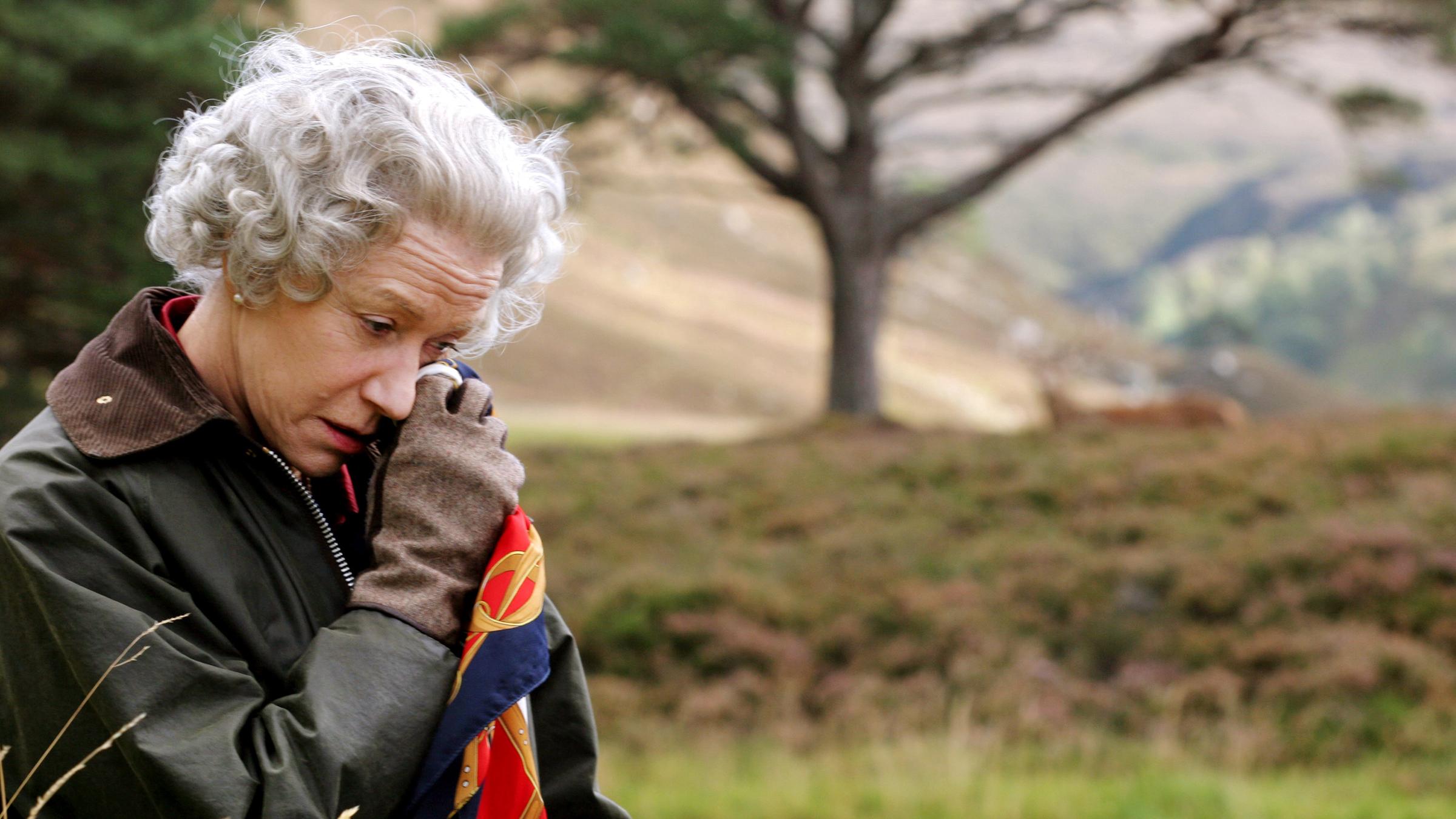 Vor dem unscharfen Hintergrund eines Baums in der Natur steht die Queen (Helen Mirren), die eine Wachsjacke mit Cordkragen und Lederhandschuhe trägt und mit gesenktem Kopf sich mit einem Seidentuch die Partie unter ihrem linken Auge betupft.