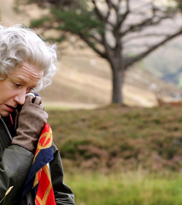 Vor dem unscharfen Hintergrund eines Baums in der Natur steht die Queen (Helen Mirren), die eine Wachsjacke mit Cordkragen und Lederhandschuhe trägt und mit gesenktem Kopf sich mit einem Seidentuch die Partie unter ihrem linken Auge betupft.