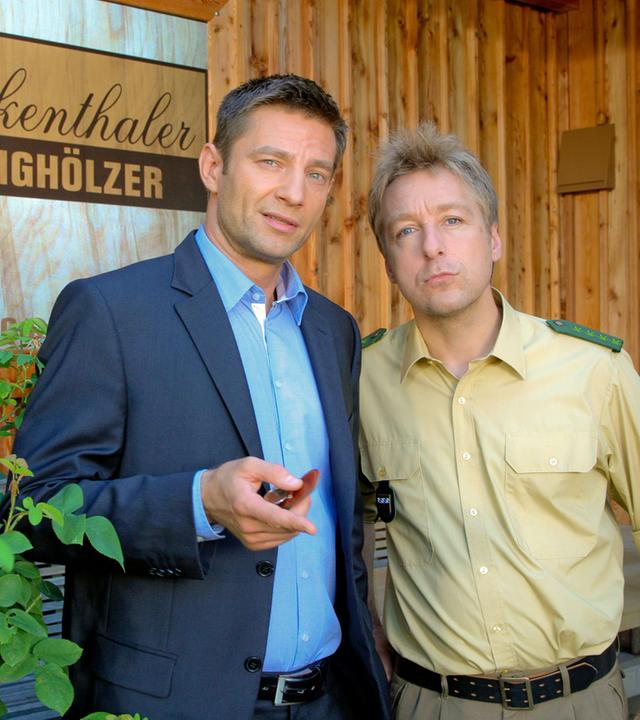 "Die Rosenheim-Cops - Die Reisen des Herrn Stuckenthaler": Kommissar Hansen (Igor Jeftic) und Polizist Michi Mohr (Max Müller) stehen vor einer Holzwand mit einem großen Firmenlogo, auf dem "Stuckenthaler Klanghölzer" steht.