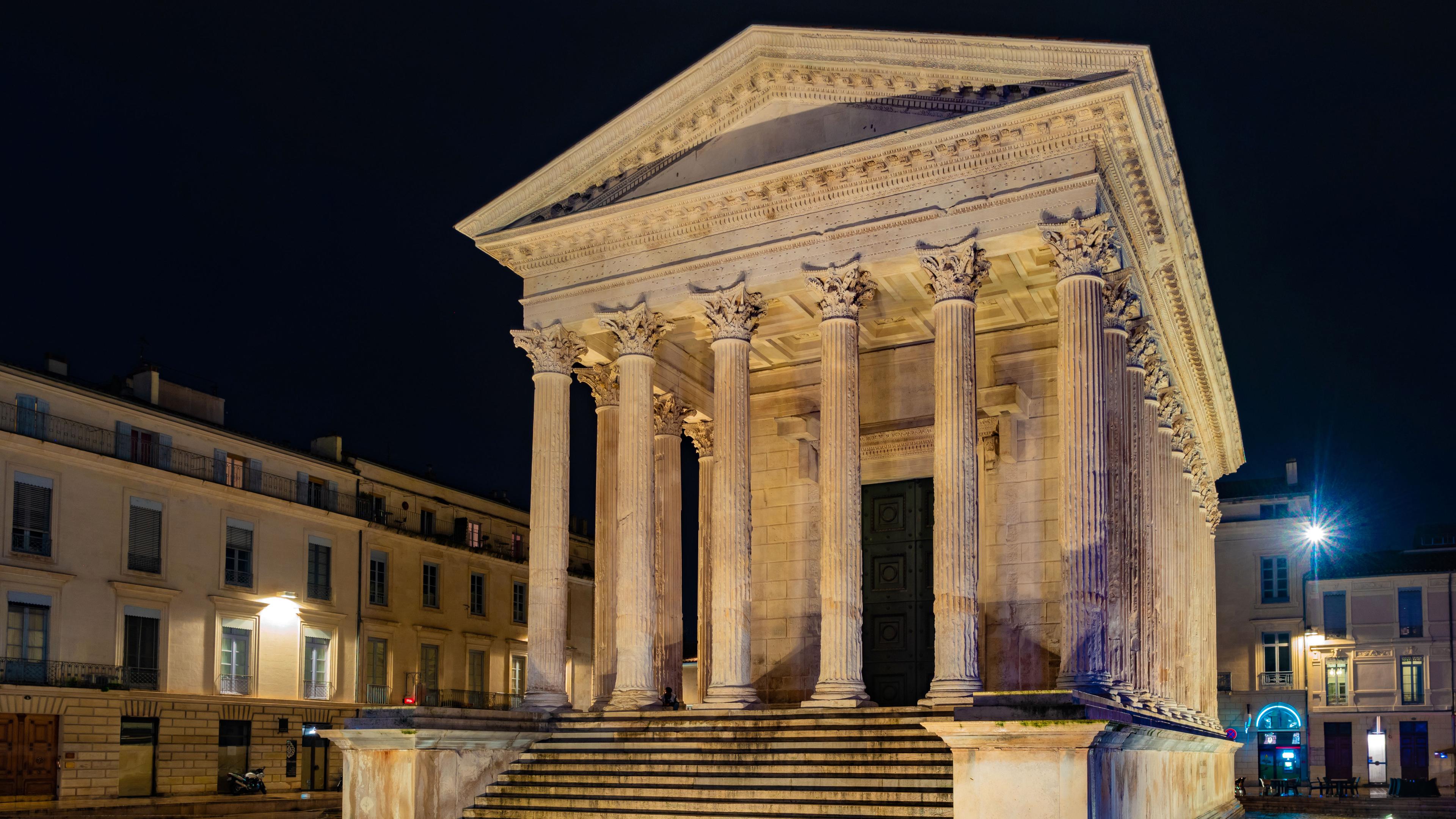 "Die Römer in Nîmes - Auf den Spuren der Antike": Maison Carrée bei Nacht: Eine Treppe führt auf ein Podium, auf dem ein Tempel mit sechs Frontsäulen und einem dreieckigen Giebel in den Himmel ragt. Das Gebäude steht auf einem freien Platz, der von Häusern umgeben ist.