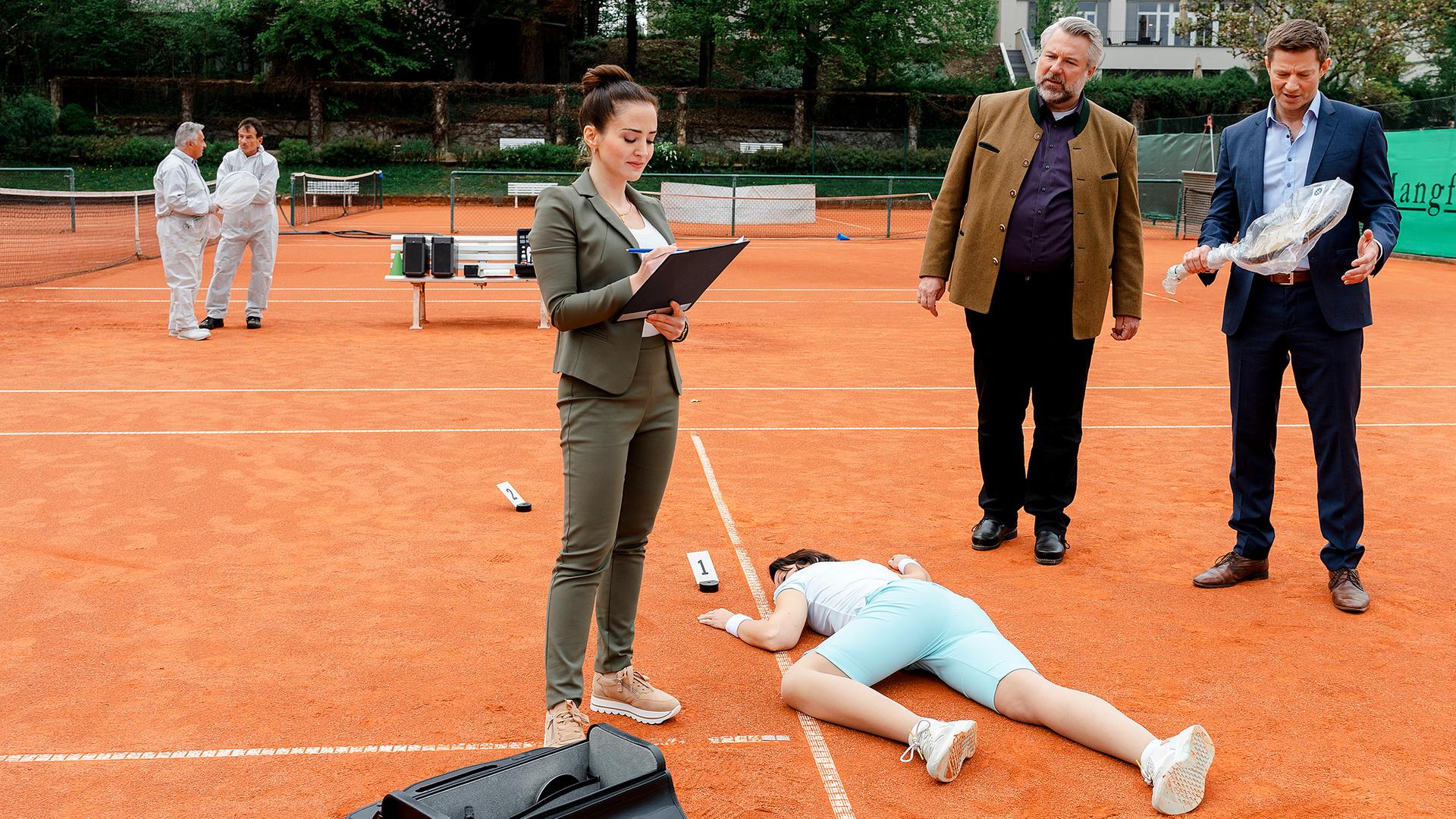 Pathologin Atay (Sevda Polat), Kommissar Stadler (Dieter Fischer) und Kommissar Hansen (Igor Jeftić) stehen um das Mordopfer herum, das bäuchlings auf dem Tennisplatz liegt. Kommissar Hansen hält einen in transparente Folie verpackten Tennisschläger in der Hand.