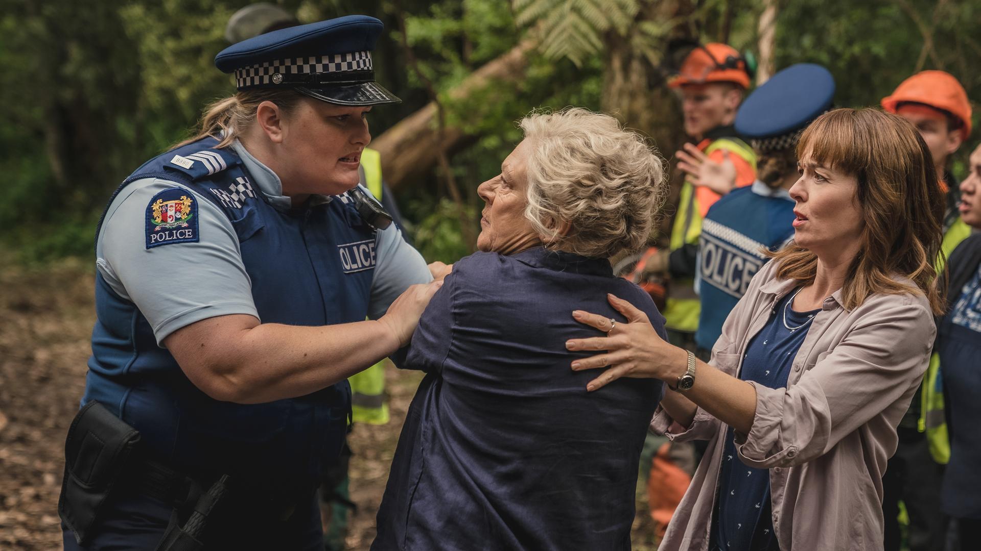 Mitch wird von einer Polizistin zurückgedrängt. Rechts daneben stehen Amanda und Miriama, die Mitch unterstützen. Im Hintergrund stehen Waldarbeiter.