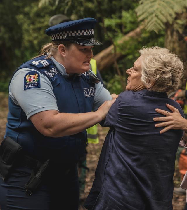 Mitch wird von einer Polizistin zurückgedrängt. Rechts daneben stehen Amanda und Miriama, die Mitch unterstützen. Im Hintergrund stehen Waldarbeiter.