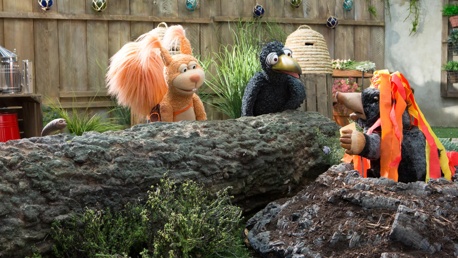 Eichhörnchen Ella und Rabe Rudi hocken vor dem Maulwurfshügel, aus dem Maulwurf Grete herausschaut. Grete trägt einen Hut mit rot-gelben Kreppbändern, die an Lavaströme erinnern.