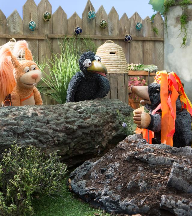 Eichhörnchen Ella und Rabe Rudi hocken vor dem Maulwurfshügel, aus dem Maulwurf Grete herausschaut. Grete trägt einen Hut mit rot-gelben Kreppbändern, die an Lavaströme erinnern.
