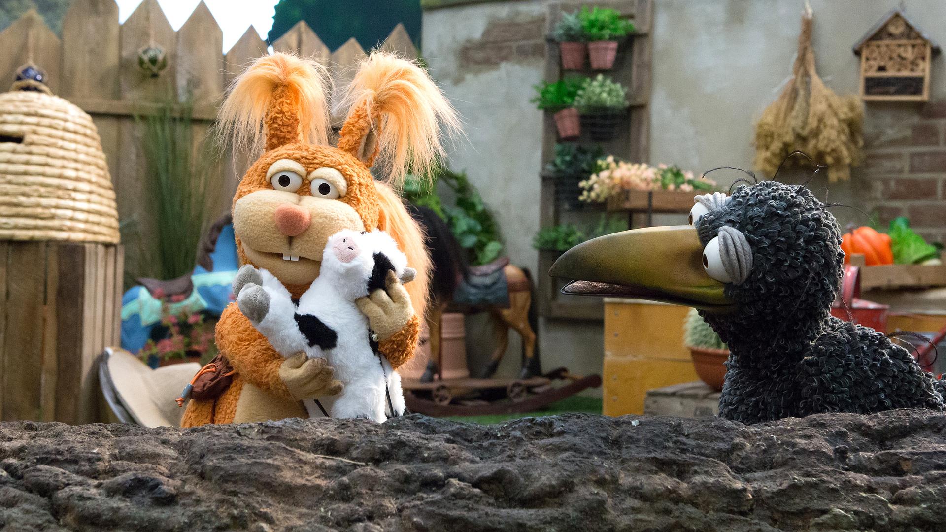 Ella und Rudi hocken auf einem Baumstamm. Rudi schaut zu Ella, die eine Plüschkuh an sich drückt.
