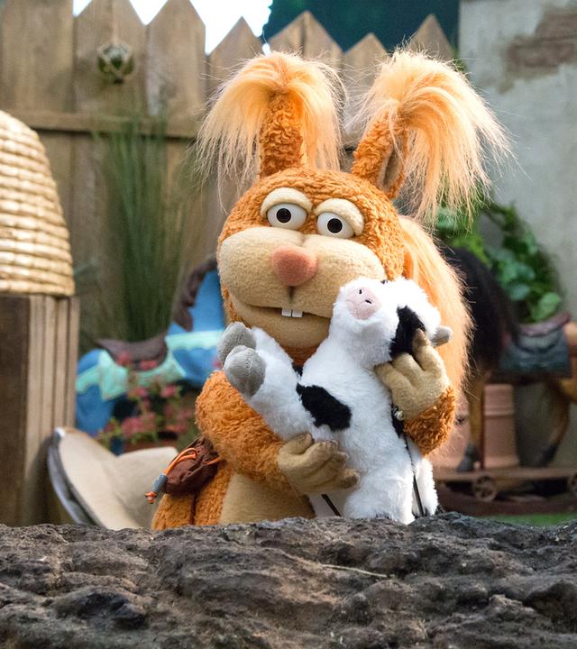 Ella und Rudi hocken auf einem Baumstamm. Rudi schaut zu Ella, die eine Plüschkuh an sich drückt.