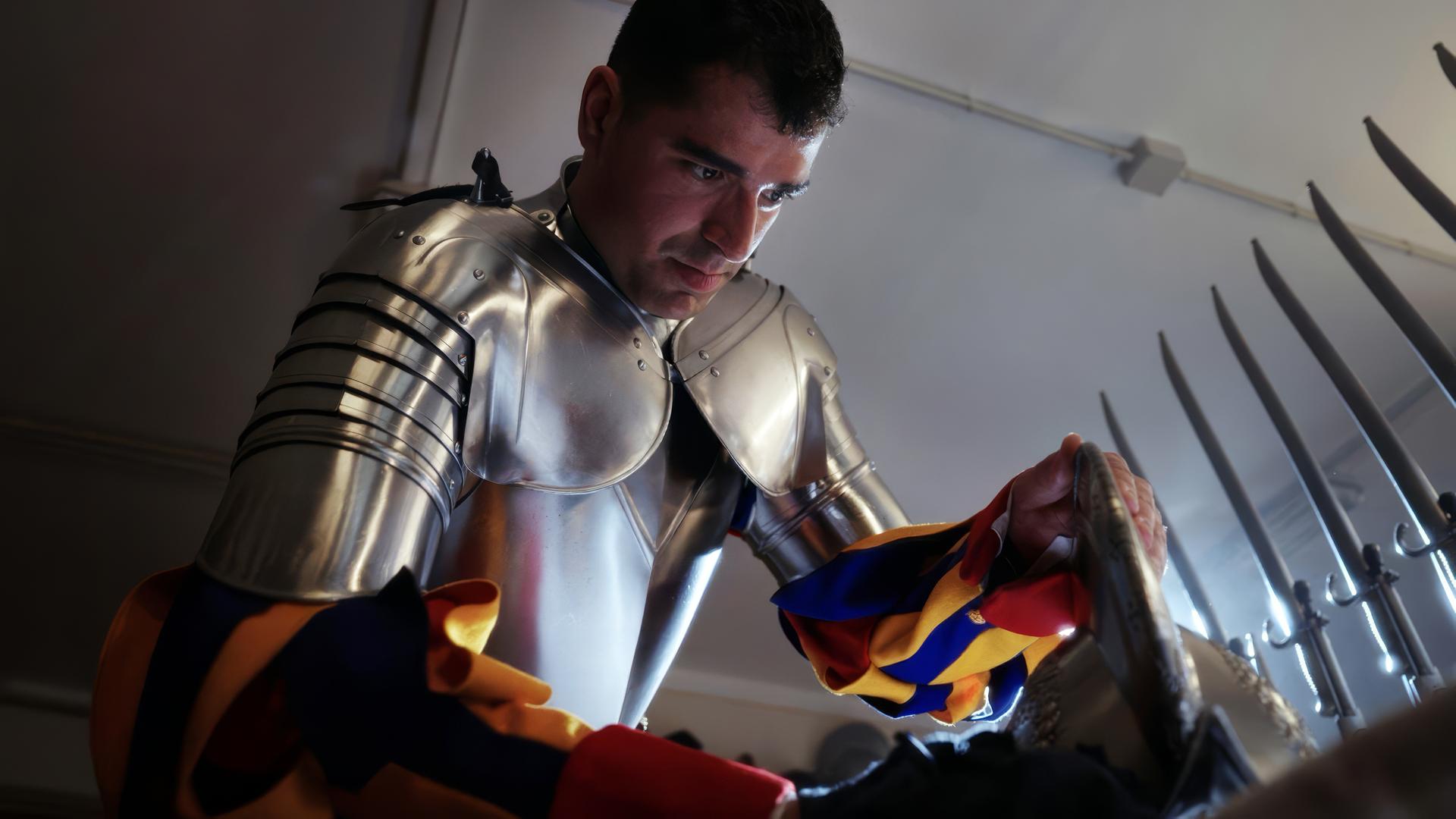 Ein Mitglied der Schweizer Garde trägt eine glänzende Rüstung und poliert ein Schild, während im Hintergrund Hellebarden zu sehen sind.